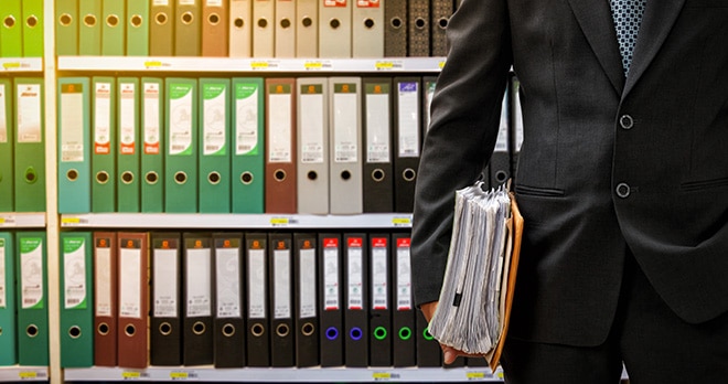 Man holding file in front of wall of shelves full of ring binders